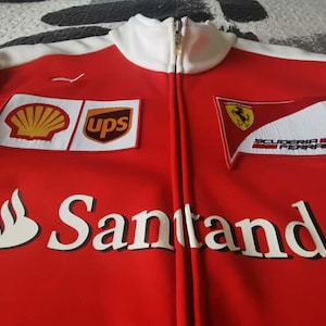 May include: Red zip-up jacket with white accents and a white collar. Features logos for Shell, UPS, and Scuderia Ferrari. The Santander logo is prominently displayed across the front.