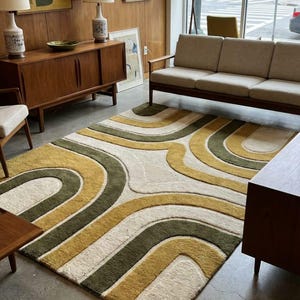 May include: A mid-century modern style rug with a cream-colored background and a repeating pattern of curved lines in olive green and mustard yellow. The rug is in a room with wood paneling, furniture, and artwork.