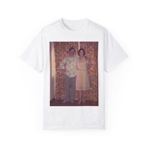 May include: A white t-shirt with a vintage photograph of a couple standing in front of a patterned curtain. The man is wearing a floral shirt and the woman is wearing a light yellow dress.