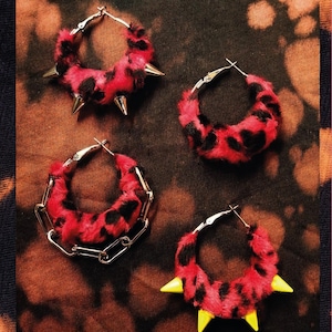 Könnte beinhalten: Vier Paar Creolen im Punkrock-Stil. Die Ohrringe haben einen rot-schwarzen Leopardenfell-Druck mit silbernen Metallreifen. Einige Ohrringe haben silberne Stacheln, andere gelbe Stacheln, und ein Paar hat eine silberne Kette.