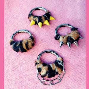 Könnte beinhalten: Vier kreisförmige Metallringe mit Leopardenmuster-Kunstfell-Akzenten. Ein Ring hat drei gelbe Stacheln, einer hat drei silberne Stacheln, einer ist schlicht und einer verfügt über eine silberne Kette, die an der Unterseite des Rings hängt.