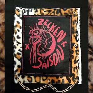 Könnte beinhalten: Ein schwarzer Stoffaufnäher mit einer pinkfarbenen Grafik einer Zecke und dem Text "ZECKEN SAISON", aufgenäht auf ein quadratisches Stück Leoparden-Kunstfell. Das Quadrat hat goldfarbene Metallösen an den Rändern und eine goldfarbene Metallkette.
