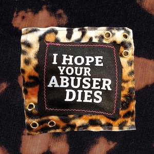 Könnte beinhalten: Ein schwarzer Stoffaufnäher mit dem weißen Text I HOPE YOUR ABUSER DIES, der auf ein Quadrat mit Leopardenmuster genäht ist. Das Quadrat mit Leopardenmuster hat goldfarbene Metallösen an den Ecken und liegt auf einem schwarzen Stoff mit braunen Bleichflecken.