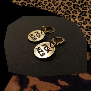 Könnte beinhalten: Ein Paar goldene Creolen mit einem runden Anhänger, auf dem in schwarzen Buchstaben "FCK NZS" steht.