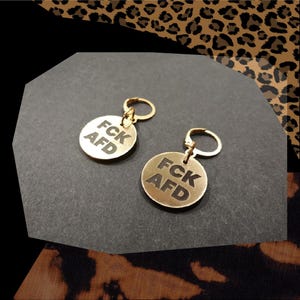 Könnte beinhalten: Zwei goldfarbene Schlüsselanhänger mit runden Metalletiketten, auf denen in schwarzer Schrift "FCK AFD" steht.