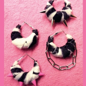 Könnte beinhalten: Vier Paar Creolen mit einem schwarz-weißen Zebrastreifenmuster. Zwei Paare haben goldfarbene Stacheln, eines hat rosa Stacheln und eines hat eine silberne Kette. Die Ohrringe sind vor einem rosa Hintergrund platziert.