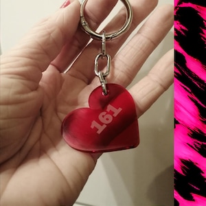 Könnte beinhalten: Ein roter herzförmiger Schlüsselanhänger mit der Aufschrift 161. Der Schlüsselanhänger ist an einem silbernen Metallring und einer Kette befestigt. Der Hintergrund ist eine Hand, die den Schlüsselanhänger hält.