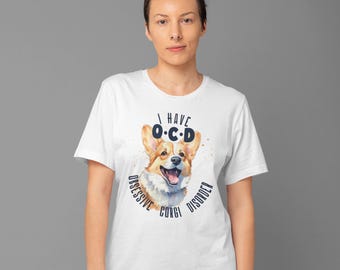 May include: White t-shirt featuring a watercolor illustration of a smiling corgi with the text "I Have O.C.D. Obsessive Corgi Disorder" in navy blue. The corgi has tan and white fur and is wearing a white t-shirt.