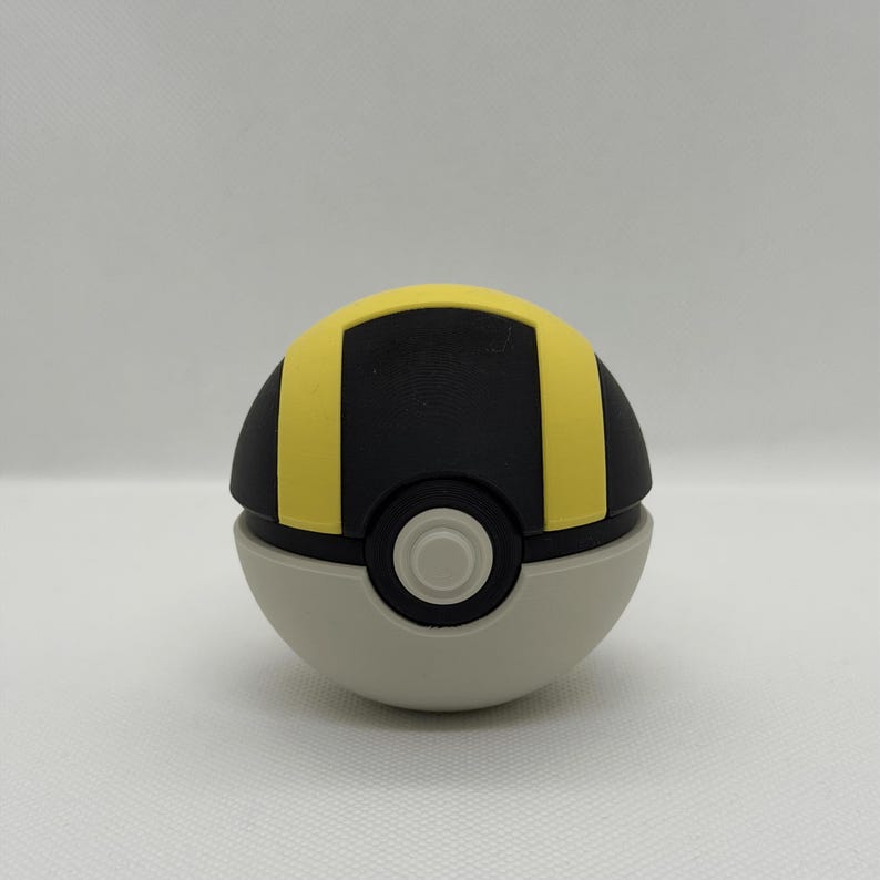 May include: A gray, black, and yellow Pok&eacute; Ball. The top half is black with a yellow stripe, and the bottom half is gray. A white circle is in the center of the ball.