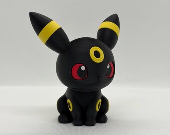 May include: A black Umbreon figurine with yellow accents on its ears and body. The figurine has red eyes and a yellow ring on its forehead. The figure is sitting on a white surface.
