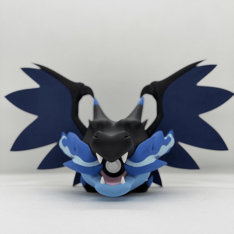 May include: A detailed sculpture of a dragon's head, viewed from the front. The dragon has a black snout and head, with blue and black wings. The dragon's mouth is open, showing white teeth and a pink interior. The sculpture is set against a white background.