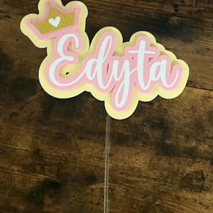 May include: A layered cake topper with the name "Edyta" in white script, outlined in pink, and set against a yellow background. A gold glitter crown with a white heart sits above the name. The topper is attached to a clear plastic stick.