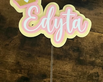 May include: A layered cake topper with the name "Edyta" in white script, outlined in pink, and set against a yellow background. A gold glitter crown with a white heart sits above the name. The topper is attached to a clear plastic stick.