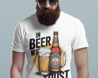May include: A white short-sleeved t-shirt featuring a graphic of a beer glass and a beer bottle. The text reads IN BEER WE TRUST. The illustration includes a golden beer with white foam and a brown bottle with a label, set against a yellow sunburst.