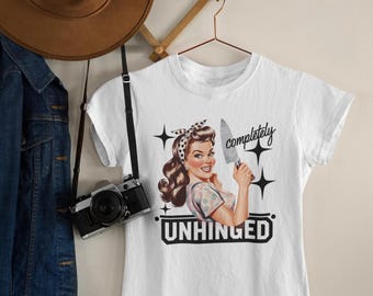 May include: White t-shirt with a retro illustration of a woman holding a knife, the text "completely unhinged" is above and below the image. A brown hat and a denim jacket hang on the wall.