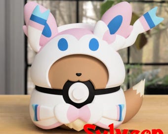 May include: A Sylveon figurine in a sitting pose. The figure is primarily white, pink, and blue, with a brown face and a fox-like tail. The word "Sylveon" is written in red at the bottom.