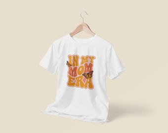 May include: White t-shirt with the text "IN MY MOM ERA" in orange and peach retro font, with two butterfly illustrations. The shirt is hanging on a wooden hanger against a light beige background.
