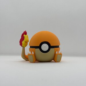 May include: An orange and tan figure resembling a Charmander, a character from the Pokémon franchise. The figure has a round orange head, a black band, and a white circle in the center. It has tan feet and a tail with a red and yellow flame.