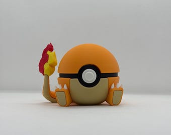 May include: A Charmander-like figure in orange and tan, a character from the Pokémon franchise. It features a round orange head, a black band, and a white circle. The figure has tan feet and a tail with a red and yellow flame.