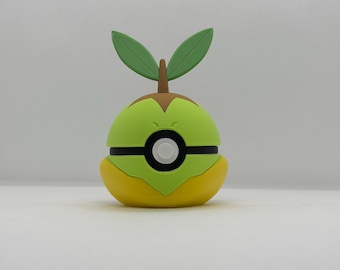 May include: A green and yellow novelty item resembling a stylized fruit or character. It features a round, segmented body with a black stripe and a white circle. Two green leaves and a brown stem sprout from the top. The item is likely a collectible or decorative piece.