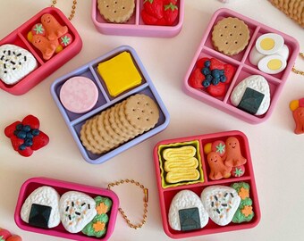 May include: Miniature food-themed trinket boxes in pink, blue, and green. Each box contains detailed replicas of snacks like crackers, strawberries, sushi, and eggs. Some boxes have gold-colored chain attachments.