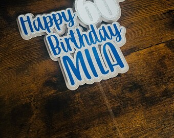 May include: A layered cake topper with the words "Happy Birthday MIA" and the number "60". The text is in a blue script font with a white outline and a silver glitter accent. The topper is attached to a clear plastic stick.
