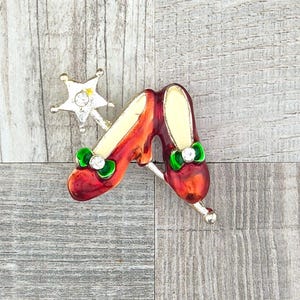 May include: A gold-tone metal brooch featuring two red shoes with green bows and clear rhinestones, accompanied by a small star-shaped wand charm with a central clear rhinestone. The design is finished with enamel detailing on the shoes and bows.