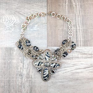 May include: A multi-strand necklace with a large, black and white zebra print pendant. The pendant is surrounded by rhinestones and the necklace is made of silver chains and black beads.