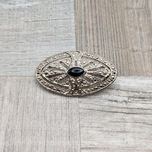 May include: A silver oval brooch with a black stone in the center. The brooch is decorated with a geometric design.