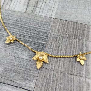 May include: A gold-tone chain necklace featuring three evenly spaced leaf-shaped charms. Each leaf charm has a textured surface and is accented with a single small white pearl at its center. The necklace is constructed with a twisted link chain.