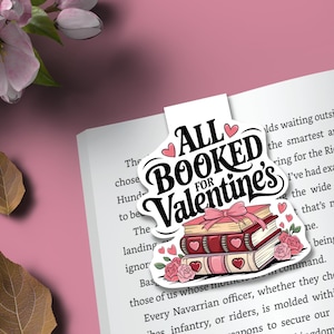 May include: A white bookmark with the text "ALL BOOKED FOR Valentine's" in a heart-shaped design. The bookmark features a stack of books with hearts and roses. The background is a pink surface with a book.