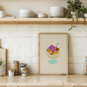 May include: A framed print of a colorful dessert in a glass bowl with a spoon. The dessert features purple ice cream, fruit, and other toppings. The print is displayed on a kitchen counter with other kitchen items.