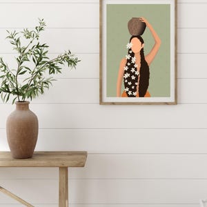May include: Framed art print featuring a stylized illustration of a person with long, dark hair adorned with white flowers, balancing a brown pot on their head. The background is a muted green, and the frame is a natural wood color.