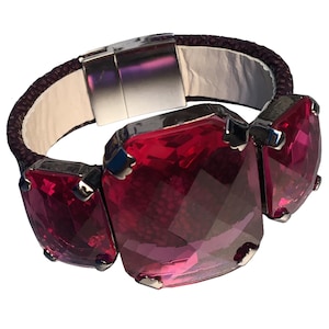 Op de afbeelding: Een armband met drie grote, facetgeslepen, robijnrode edelstenen, gezet in zilverkleurig metaal. De armband heeft een donkerpaarse gevlochten leren band met een zilveren sluiting en een witte leren achterkant.