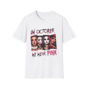 Op de afbeelding: Een wit t-shirt met korte mouwen en een grafische print van vier gestileerde gezichten met horror-make-up en maskers. De tekst boven de afbeeldingen luidt IN OCTOBER en de tekst eronder luidt WE WEAR PINK in een verweerd, donkerrood lettertype.