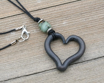 Peut inclure: Un collier en cordon noir avec un fermoir argenté et un pendentif en forme de cœur en métal. Le pendentif est attaché à une perle de pierre verte avec des accents argentés.
