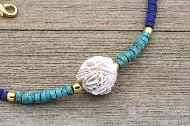 Puede incluir: Un collar de cuentas de coral blanco con cuentas turquesa y azules. El collar tiene un cierre dorado.