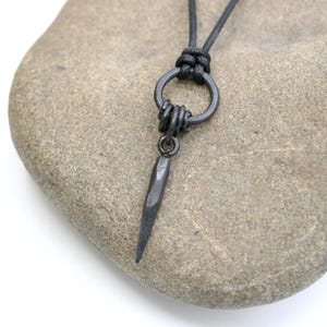 May include: A dark grey metal pendant necklace with a spike-shaped charm. The pendant hangs from a black cord, knotted at the top. The necklace is displayed on a light brown stone against a white background.