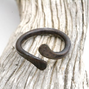 May include: A dark brown metal ring with a curved design. The ring is open-ended, with one side curving inward. The ring is set against a textured, light-colored wooden surface.