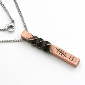 May include: A silver chain necklace with a copper pendant. The pendant is a twisted bar with black accents and a symbol engraved on it.