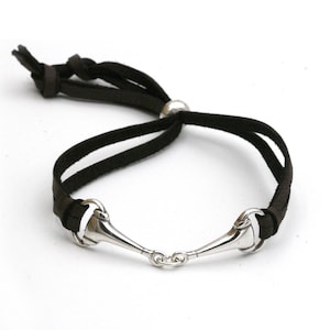 May include: Black leather bracelet featuring a silver-toned equestrian bit design. The bracelet has an adjustable sliding knot closure and a small silver bead. A stylish accessory for equestrian enthusiasts.