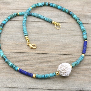 Puede incluir: Un collar de cuentas turquesa y azul con un colgante de concha blanca. El collar est&aacute; hecho con cuentas peque&ntilde;as y redondas y tiene un cierre dorado.