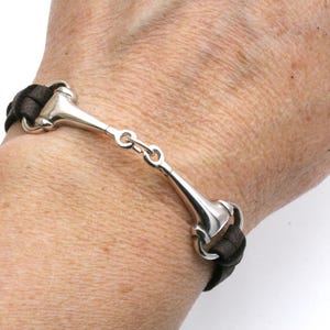 May include: A silver-toned bracelet with a horse bit design. The bracelet features a central bit connected to dark brown leather straps. The design is a classic equestrian style, suitable for everyday wear.