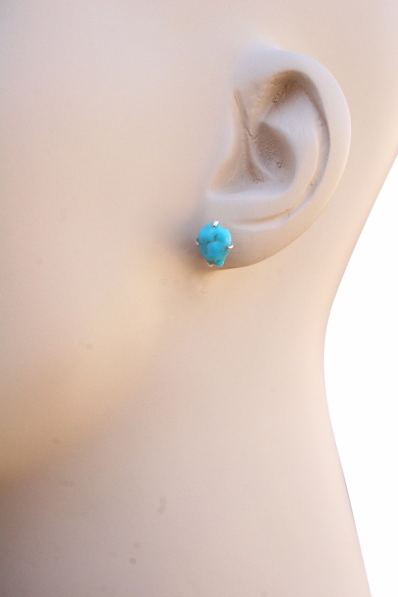May include: A pair of turquoise stud earrings with silver settings. The earrings are shown on a mannequin's ear.