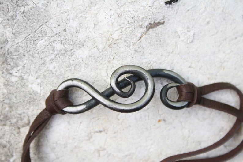 May include: A brown leather cord bracelet with a silver metal infinity symbol charm. The symbol is twisted and has a swirl design.