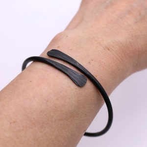 May include: A black cuff bracelet with a textured finish. The bracelet wraps around the wrist with two decorative, flared ends. The jewelry is shown on a person's arm against a white background.