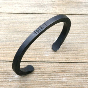 May include: A black metal cuff bracelet with a minimalist design. The open-ended bracelet features a series of etched lines near the top. The bracelet is shown on a light wood surface.