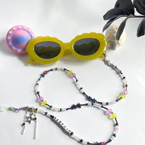 May include: A yellow plastic glasses chain with a beaded design featuring pink, green, and iridescent star-shaped beads. The chain has a silver charm with two stars and the name "SEOKJIN" spelled out in white beads.