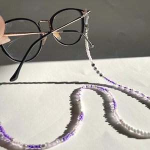 Può includere: Una catena per occhiali in perle bianche e viola con la parola "BTS" scritta in perle bianche.