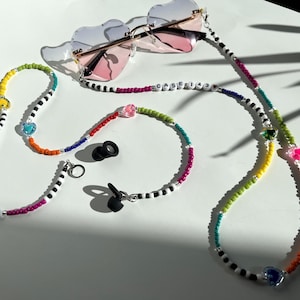 May include: A pair of heart-shaped sunglasses with a gold frame and pink lenses rests above a beaded eyeglass chain. The chain is multi-colored with the text "CASE143" and heart-shaped beads. Also included is a matching beaded bracelet.